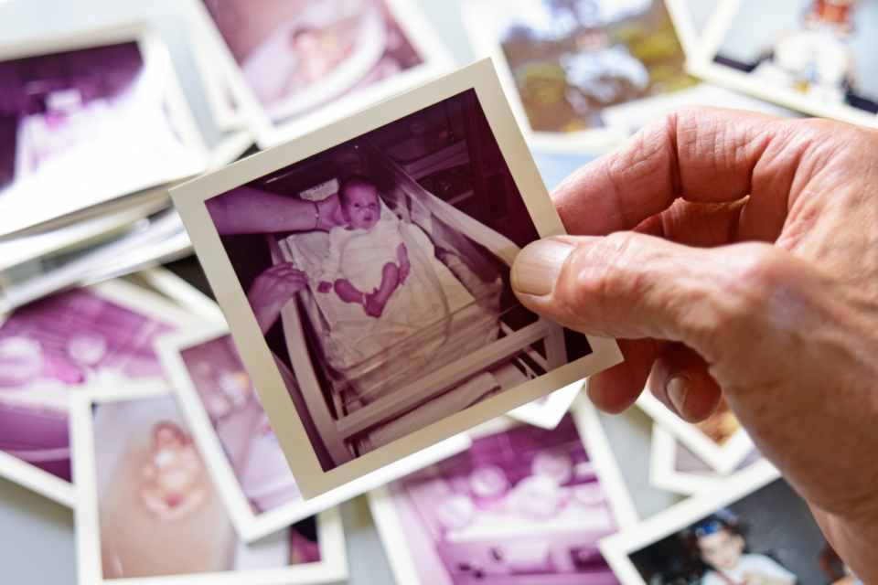 An older hand holding a photo of a baby. The photo is polaroid, looks dated and is of a baby in hospital after birth. There are blurred photos scattered in the background.