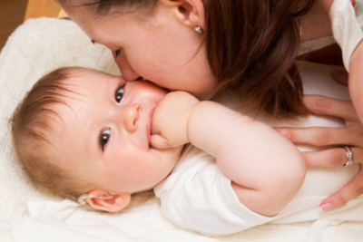 A baby is laid on a changing mat. Their mum is kissing their cheek, and the baby is looking directly at the camera, smiling with their hand in their mouth. They have blue eyes, light skin and hair and a hearing aid in their right ear.