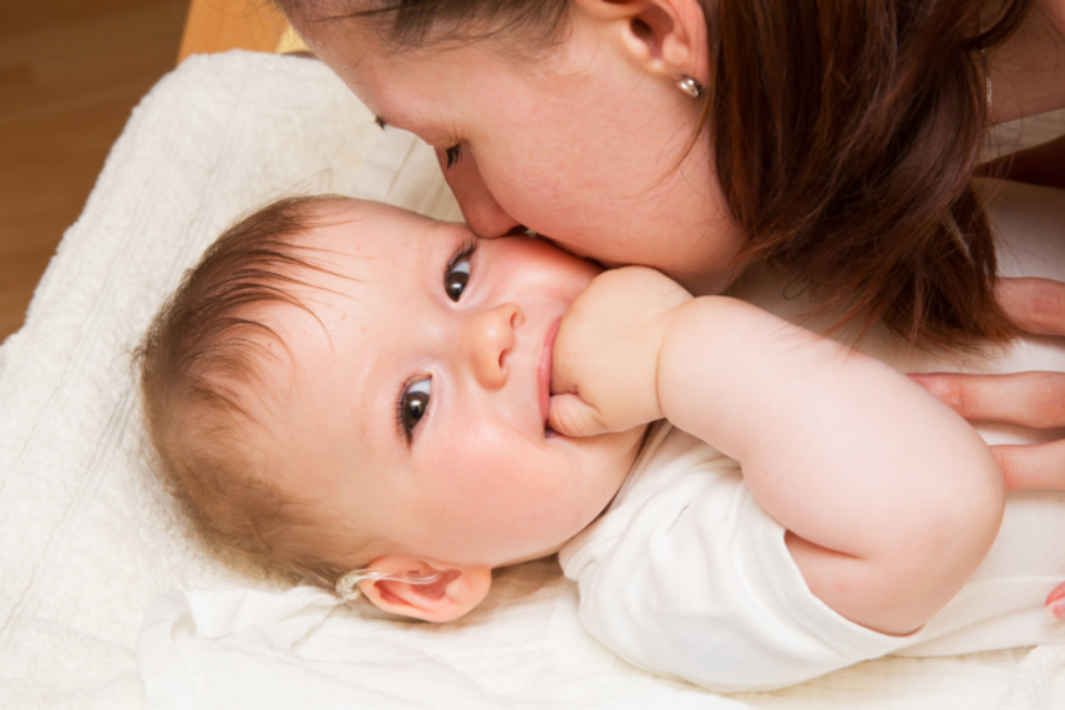 A baby is laid on a changing mat. Their mum is kissing their cheek, and the baby is looking directly at the camera, smiling with their hand in their mouth. They have blue eyes, light skin and hair and a hearing aid in their right ear.
