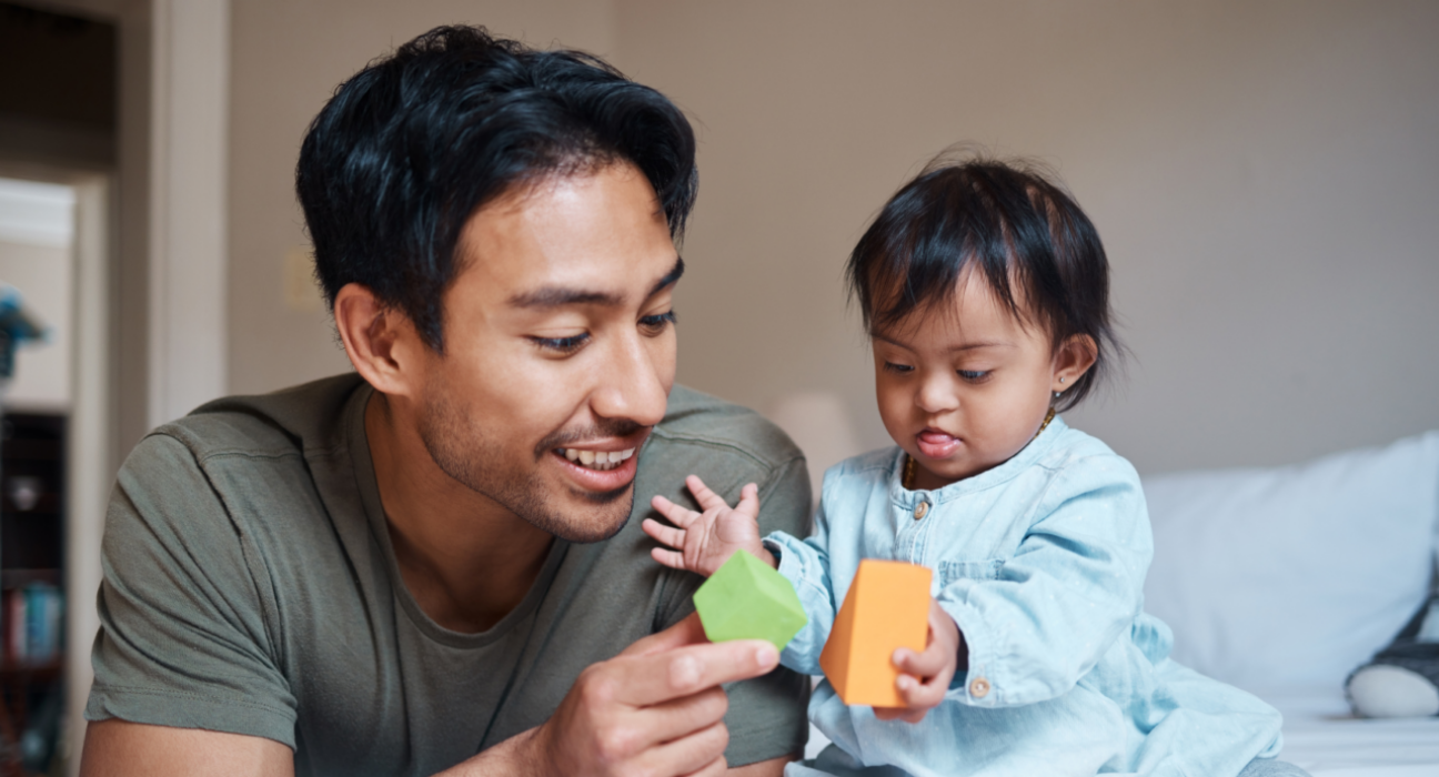 A dad and baby are playing together with colourful blocks. Dad is smiling and the baby's tongue is slightly stuck out in concentration. The baby. Both have dark hair and a brown skin tone, and the baby wears a blue top.