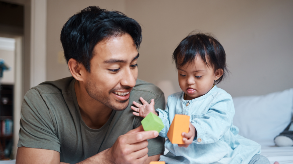 A dad and baby are playing together with colourful blocks. Dad is smiling and the baby's tongue is slightly stuck out in concentration. The baby. Both have dark hair and a brown skin tone, and the baby wears a blue top.