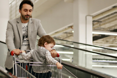A dad and a baby are in a shop with a shopping trolley. The baby is a young toddler and sits in the trolley seat. The dad is looking down at him. They both have light skin and the baby has light brown hair, and the dad darker brown hair with a beard.