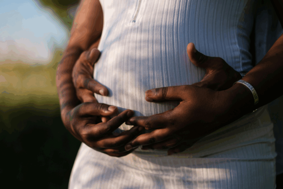 Two hands are holding a pregnant stomach. Both hands have a dark skin tone and hold the stomach which has a white dress over the bump. The couple seem to be outside and the lighting is golden.