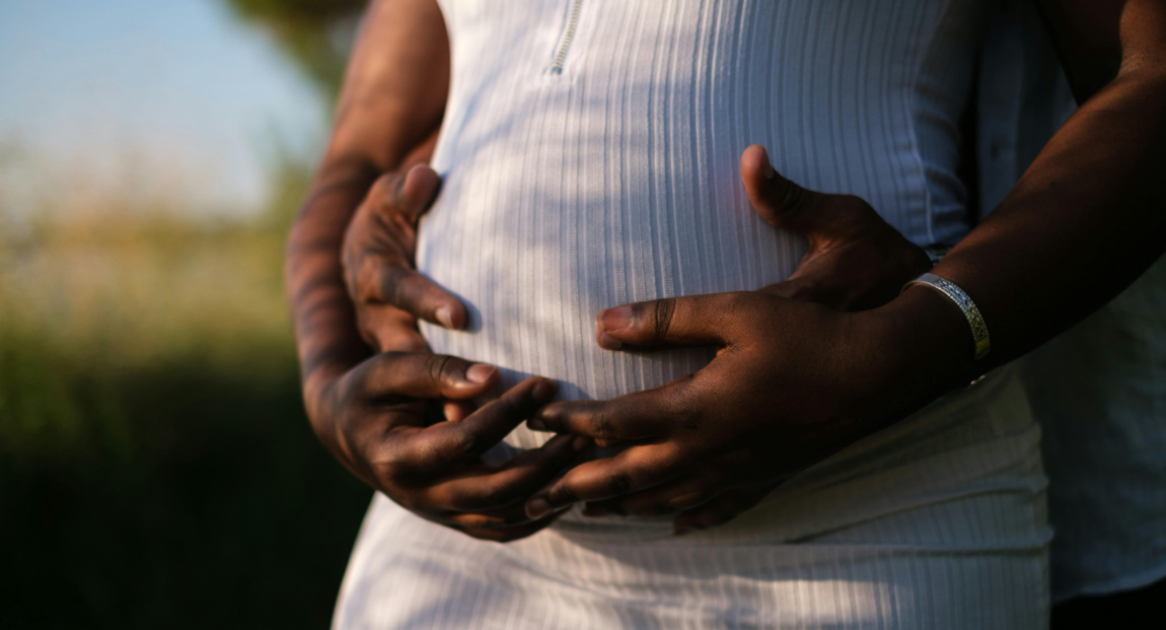 Two hands are holding a pregnant stomach. Both hands have a dark skin tone and hold the stomach which has a white dress over the bump. The couple seem to be outside and the lighting is golden.