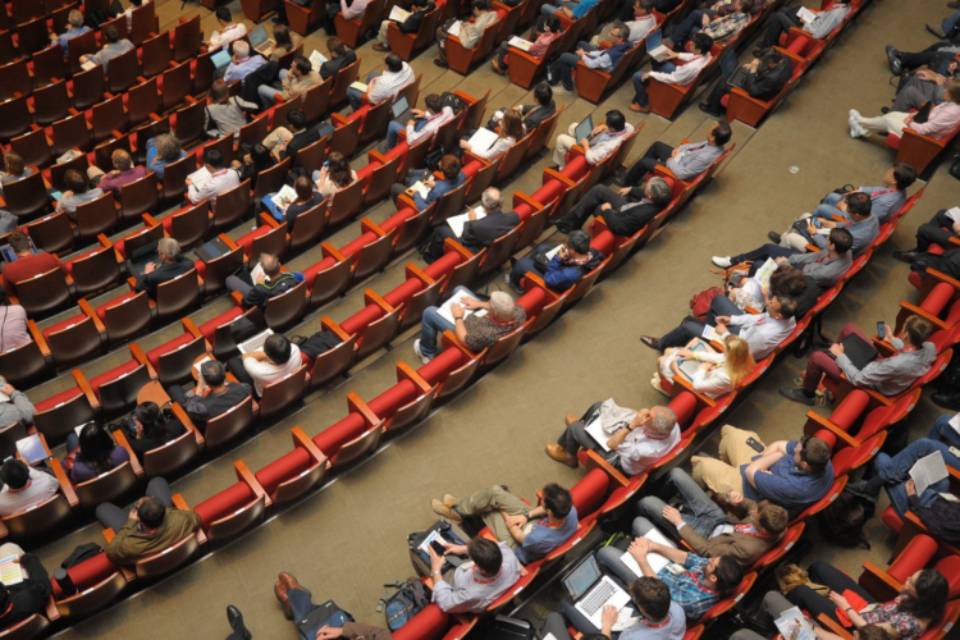 A shot from the top of an auditorium looking down at hundreds of people sat in seats at a conference.