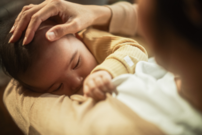 A baby is breastfeeding. They are clutching onto their mother's t-shirt and wear a light yellow top. The mum has the hand gently placed on the baby's head.