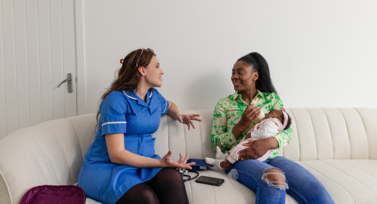 Mother bottle-feeding a baby, sitting on a sofa beside a health professional who is talking to her.