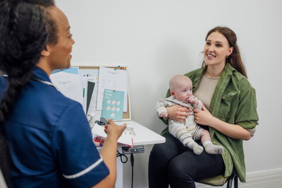Mother sitting on a chair in a doctor's office with a baby on her lap, talking to a health professional.
