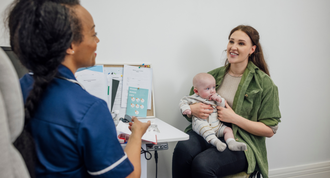 Mother sitting on a chair in a doctor's office with a baby on her lap, talking to a health professional.