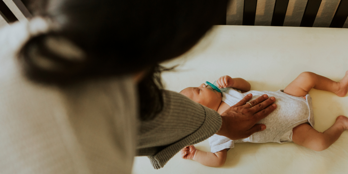 A baby is sleeping on their back in their cot with a dummy in their mouth. They have a medium skin tone and their mum has her hand on their chest.
