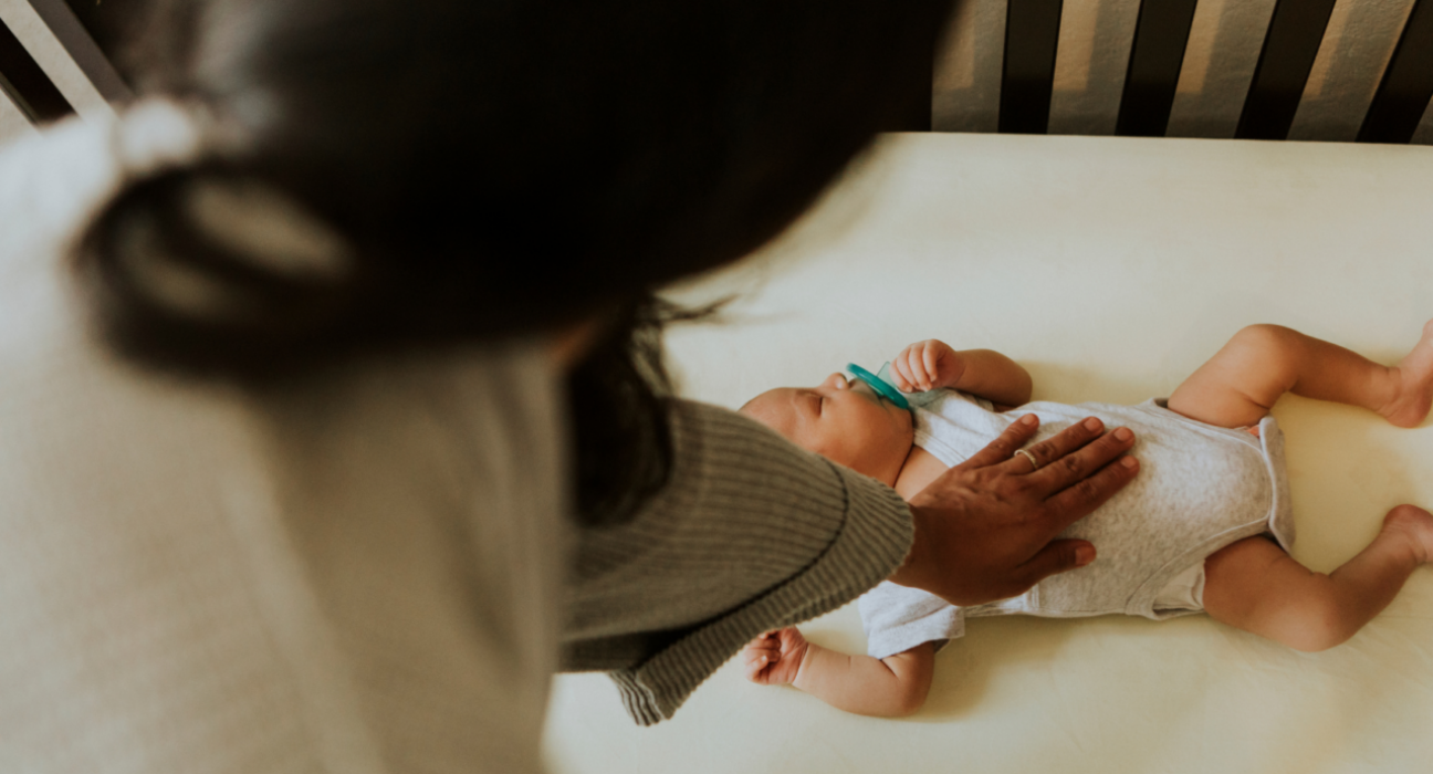 A baby is sleeping on their back in their cot with a dummy in their mouth. They have a medium skin tone and their mum has her hand on their chest.