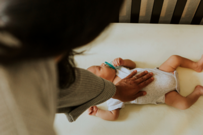 A baby is sleeping on their back in their cot with a dummy in their mouth. They have a medium skin tone and their mum has her hand on their chest.
