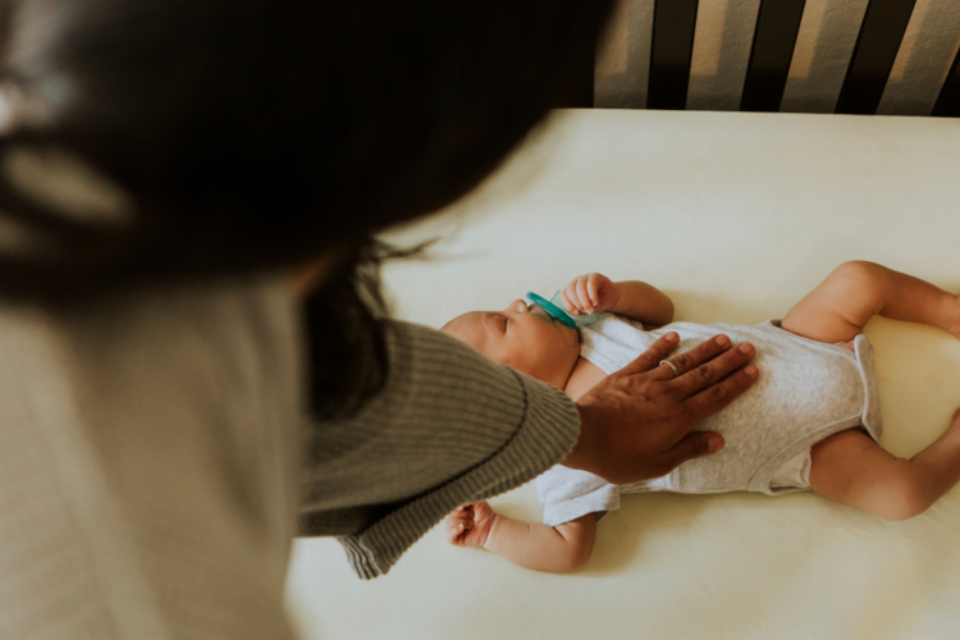A baby is sleeping on their back in their cot with a dummy in their mouth. They have a medium skin tone and their mum has her hand on their chest.
