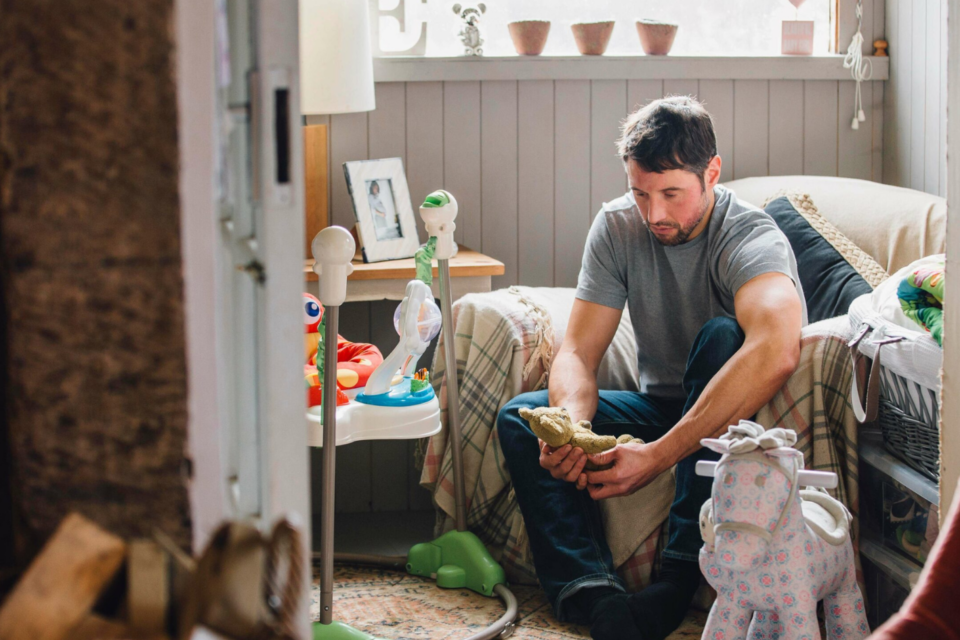 Man sitting on an armchair in a child's bedroom amongst toys, looking solemnly at a teddy bear in his hands.