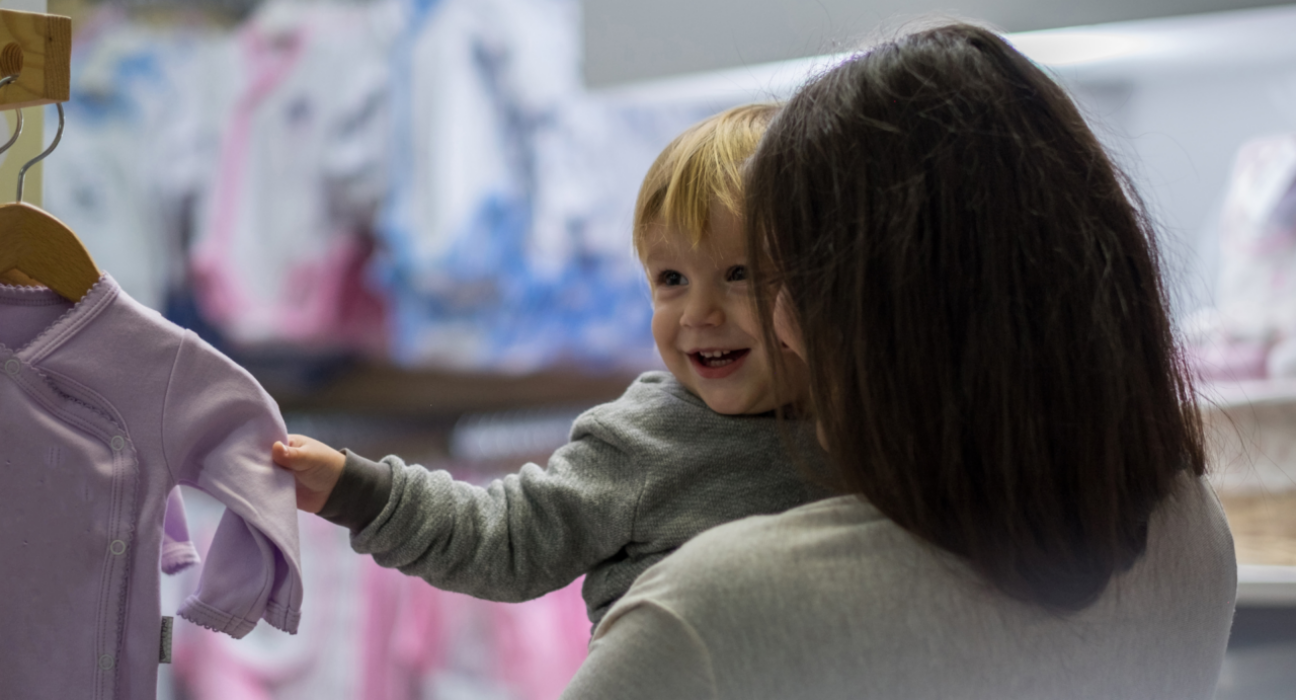 Baby and a mum are shopping together. The baby is smiling at someone out of frame and the mum is looking at the baby. The baby has blonde hair, light skin and is grasping onto a baby grow.