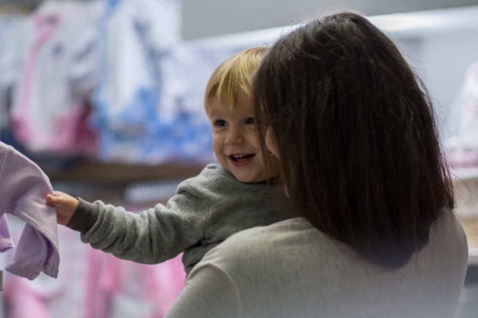 Baby and a mum are shopping together. The baby is smiling at someone out of frame and the mum is looking at the baby. The baby has blonde hair, light skin and is grasping onto a baby grow.