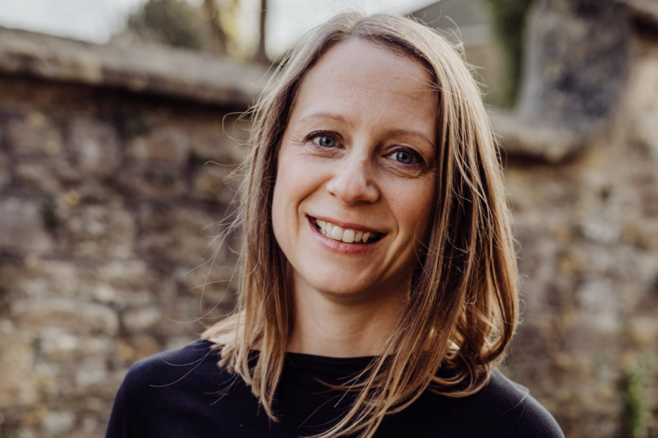 Portrait of Anna Pease wearing a black top, smiling at the camera in front of a brick wall.