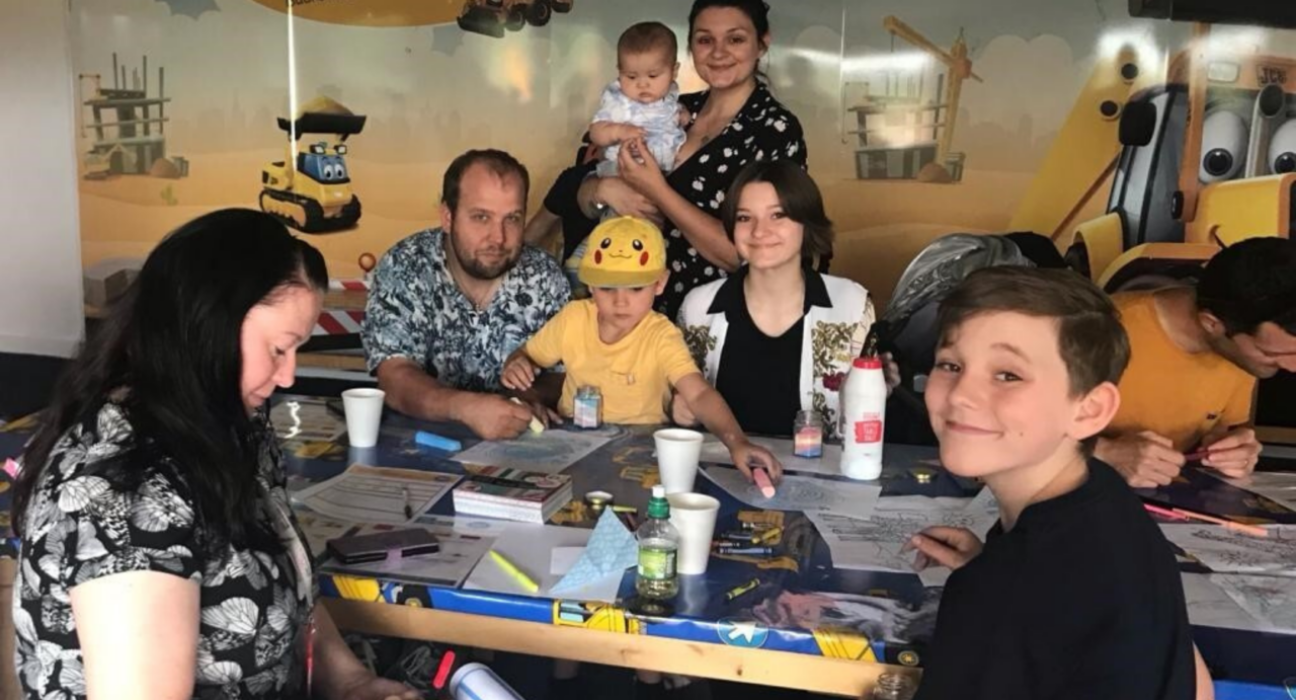 Family sitting at a table at a family day, doing colouring in and smiling at the camera.