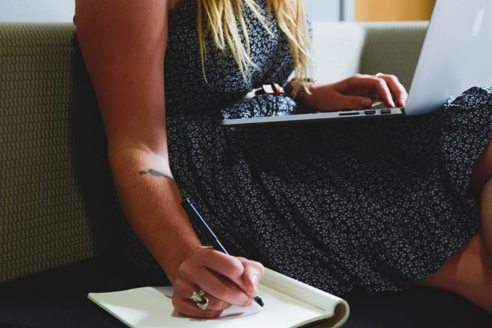 Lady witting on a chair, with a laptop on her lap, and writing in a notepad beside her.
