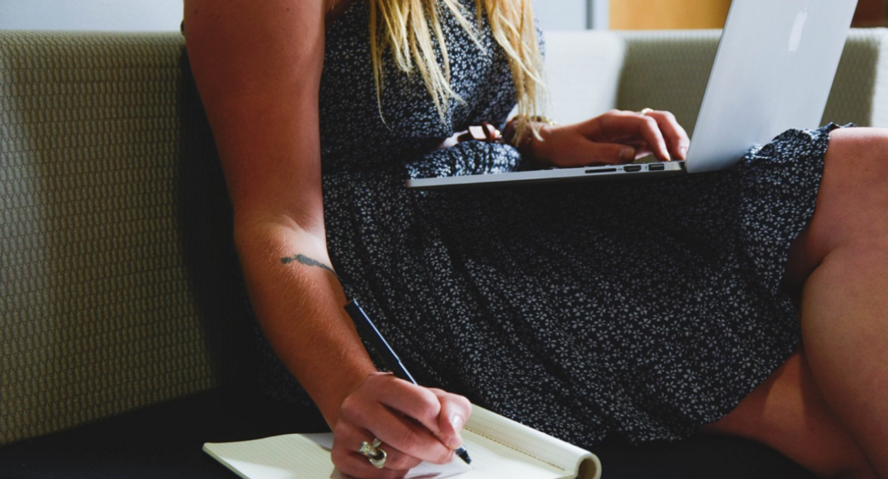 Lady witting on a chair, with a laptop on her lap, and writing in a notepad beside her.