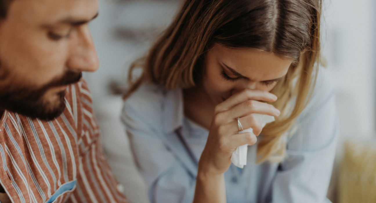 Upset lady with tissue in her hand looking down, with man sat beside her.