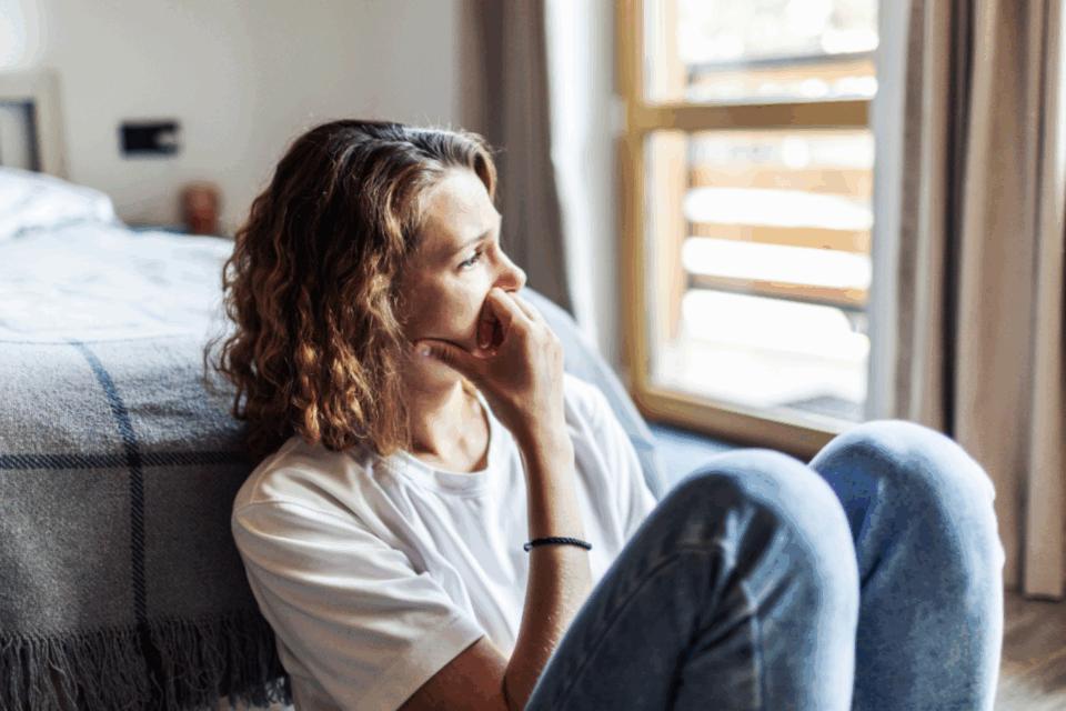 Lady sitting on the floor at the end of her bed, looking out towards the window sadly.