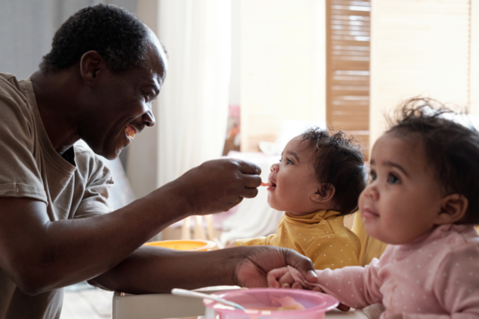 Two twins babies are being fed by their dad. They have dark skin and ruffled, dark hair. One wears yellow, the other pink and the baby in yellow is having a spoon of liquid food put into their mouth by the dad.