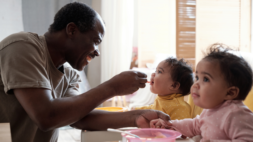 Two twins babies are being fed by their dad. They have dark skin and ruffled, dark hair. One wears yellow, the other pink and the baby in yellow is having a spoon of liquid food put into their mouth by the dad.