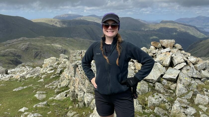 A woman in black sportswear and sunglasses, stands proudly in front of a view of the Lake District