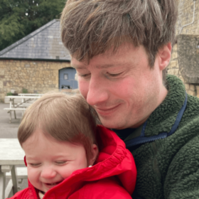Matt and baby Sam are sat on a bench. Sam is grinning cheekily and Matt is holding him, smiling and looking down at baby Sam's hands.