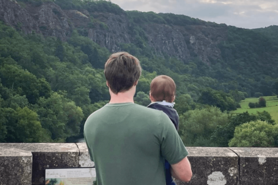 Baby Sam is being held by Matt's right arm. They are both looking out over a bridge at a beautiful, green view. Their backs are to the camera.