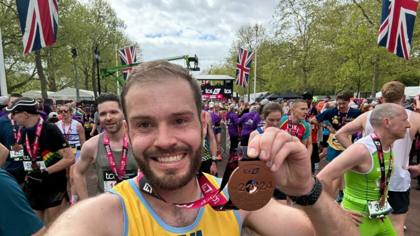 Man standing in crowd of runners smiles to camera and holds up a London marathon medal