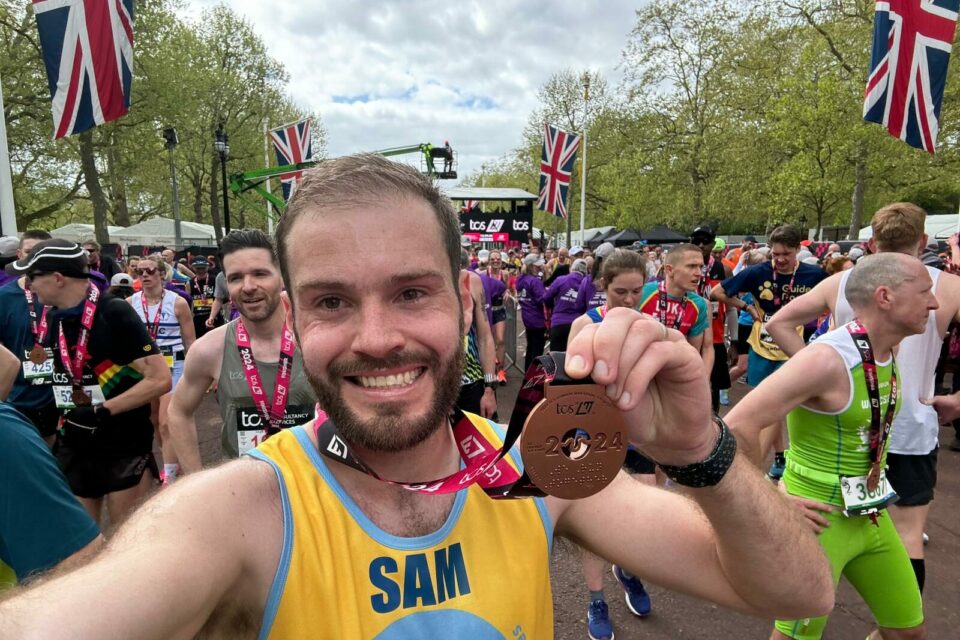 Man standing in crowd of runners smiles to camera and holds up a London marathon medal