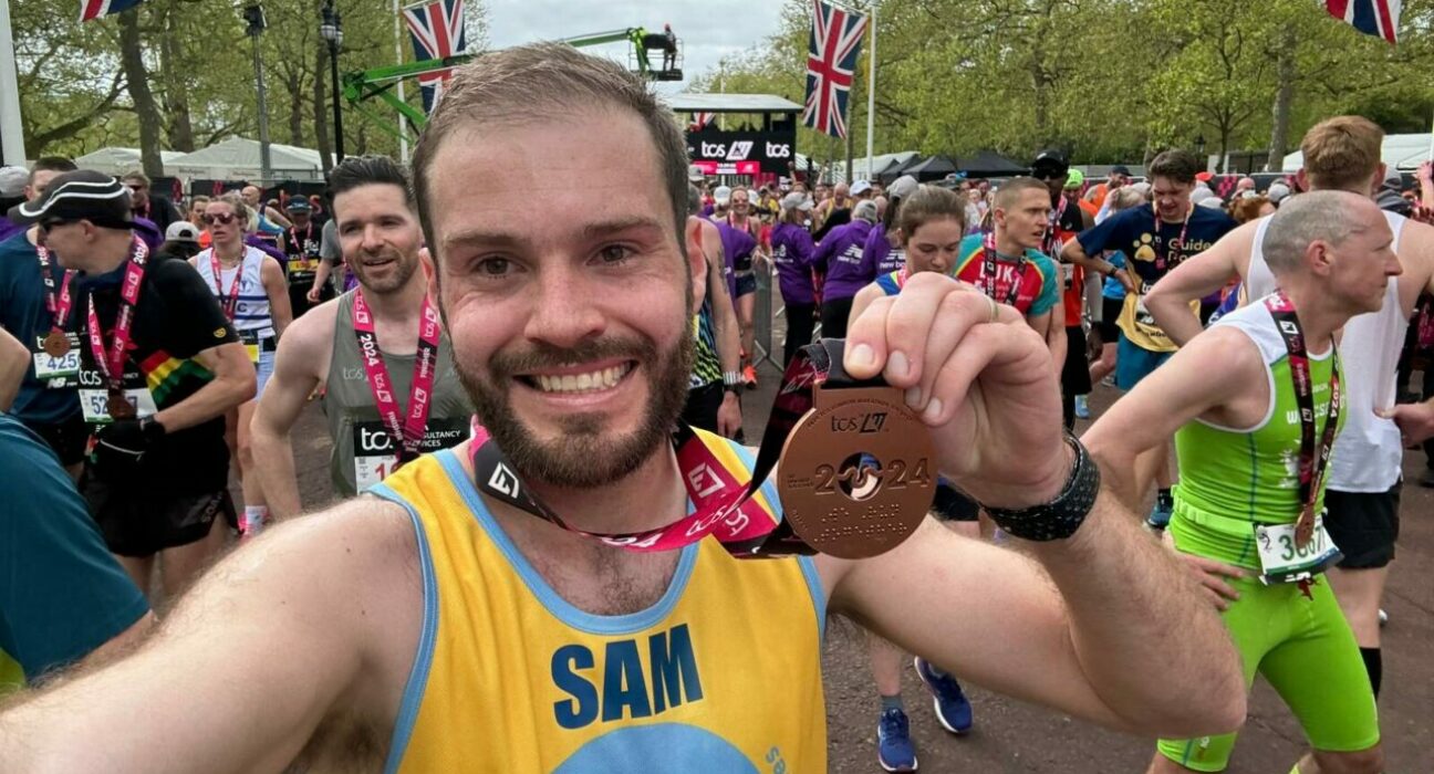 Man standing in crowd of runners smiles to camera and holds up a London marathon medal