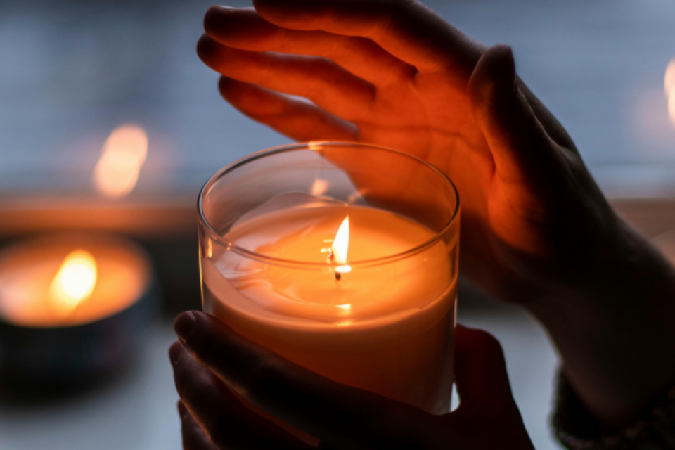 A person has lit a candle and they are holding it in one hand, and holding their other hand above the candle which is in a glass jar. In the blurred background, you can see another candle on a windowsil.