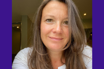 Claire is taking a selfie, looking directly into the camera lens. She is smiling, wearing a white blouse. She has brown eyes and brown hair which is curling inwards by her chin.