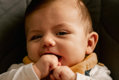 A baby is sat in a car seat. The close-up of their face shows their slight grin as they are putting their fisted hand in their mouth. They have light skin, light brown hair.