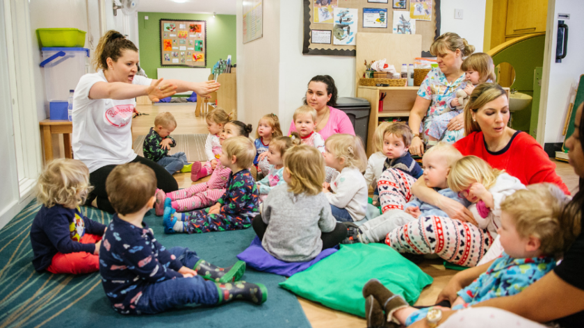 Children wearing pyjamas sit on the carpet in their nursery, watching their teacher