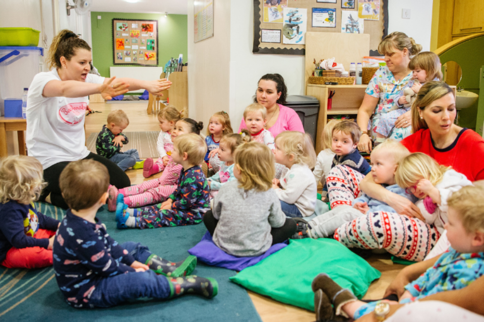 Children wearing pyjamas sit on the carpet in their nursery, watching their teacher