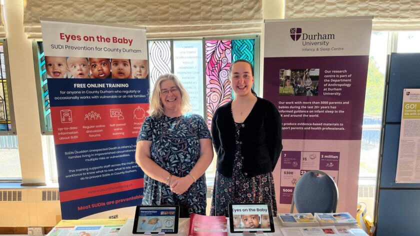 Two researchers are pictured next to one another, both women and both stood proudly. Behind them are pull up banners relating to SIDs and SUDI, and in front of them is a table full of useful information on their research projects.