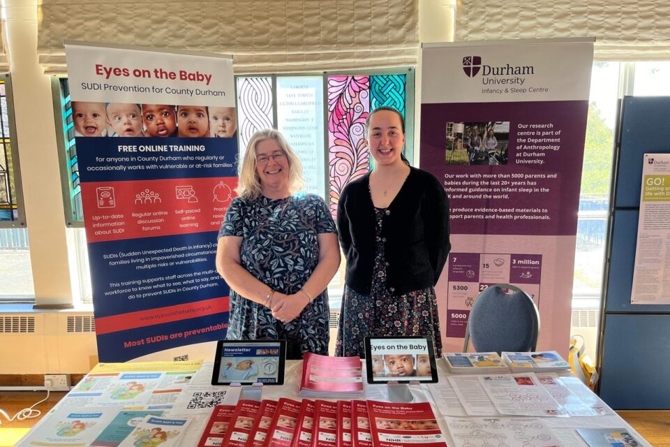 Two researchers are pictured next to one another, both women and both stood proudly. Behind them are pull up banners relating to SIDs and SUDI, and in front of them is a table full of useful information on their research projects.