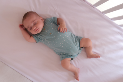Baby sleeping in a cot on their back. They have a light sleep suit on and the light is coming through the bars of the cot. They have light skin.