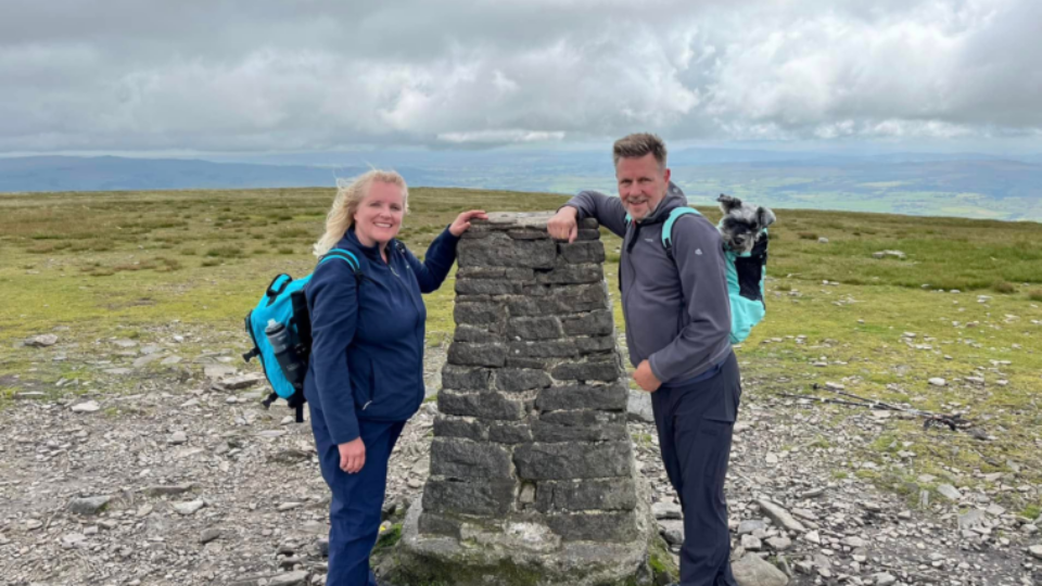 Louise and Jim are on a hike and are both leaning on a stone. Their dog, Lulu, is in a backpack that Jim is wearing. They are smiling.