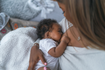 A baby is being breastfed by their mum. They look very young, have curly dark hair and dark skin. Their mum is looking down at them.