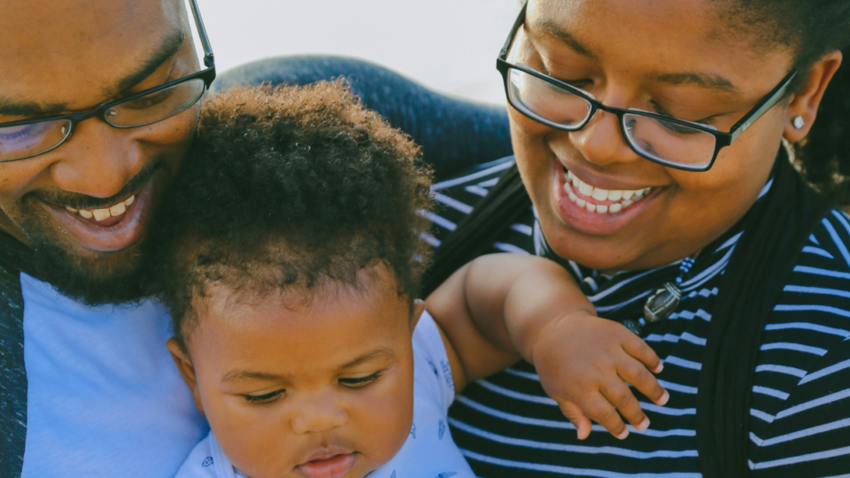 A family are pictured together close-up. They have dark skin and both mum and dad are smiling down at their baby. Their baby has the same skin and curly, dark brown hair and is looking down at something.