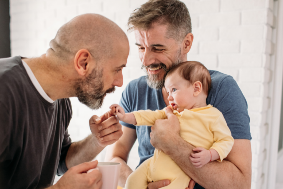One man is holding a baby with the baby's head near his chin. The other man is holding the baby's hand in his, and putting his mouth to the baby's hand. Both men are smiling.