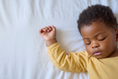 A baby is sleeping on their back. They have a yellow sleepsuit on and they have dark skin with curly hair.