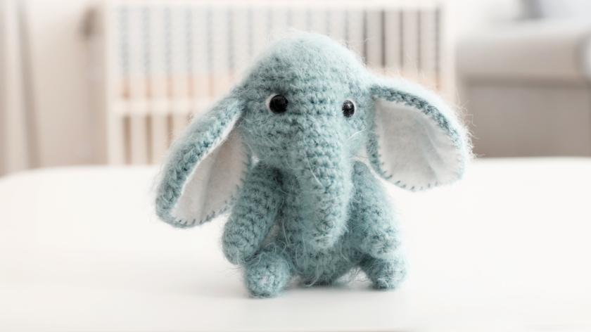 A crochet small baby toy elephant sits in front of a baby's crib