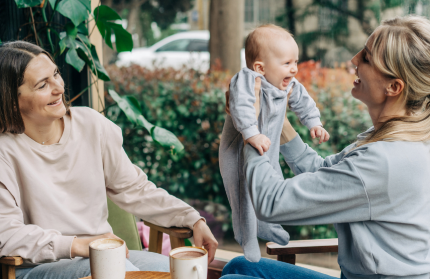 Two women are sat at a cafe and one is holding the baby up in their arms. They are both smiling, but the baby has the biggest and widest smile of them all. They have light skin and the baby is wearing a baby grow.