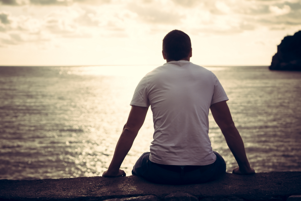 Man is sitting looking out at the sea. He has a muscular silhouette, shaved head and is slumped slightly.